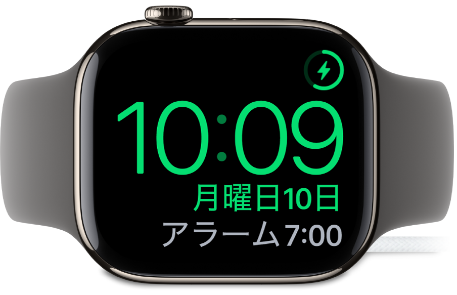 横向きに置かれ、充電器に接続されているApple Watch。画面には、右上隅に充電中のマーク、その下に現在時刻と、次のアラーム時刻が表示されています。