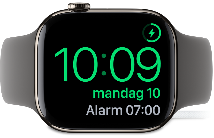 En Apple Watch som ligger på siden og er koblet til laderen, med skjermen som viser ladesymbolet øverst til høyre, gjeldende klokkeslett og tidspunktet for neste alarm.