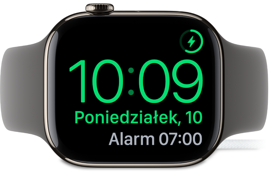Apple Watch ustawiony na boku i podłączony do ładowarki. W prawym górnym rogu ekranu widoczny jest symbol ładowania, niżej wyświetlana jest bieżąca godzina, a dalej znajduje się czas najbliższego alarmu.