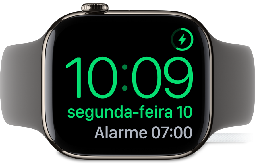 Um Apple Watch colocado de lado e ligado ao carregador, com o ecrã, com o símbolo de carregamento no canto superior direito e, por baixo, a hora atual e a hora do próximo alarme.