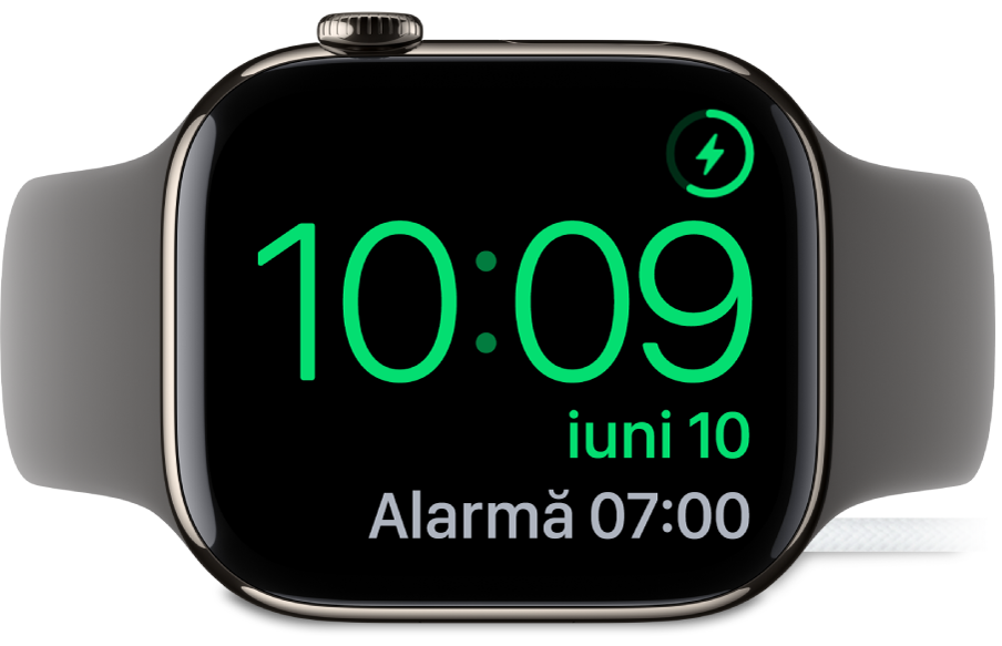 Un Apple Watch așezat pe muchie și conectat la încărcător, pe ecran fiind afișat simbolul de încărcare în colțul din dreapta sus, ora actuală dedesubt și ora alarmei următoare.