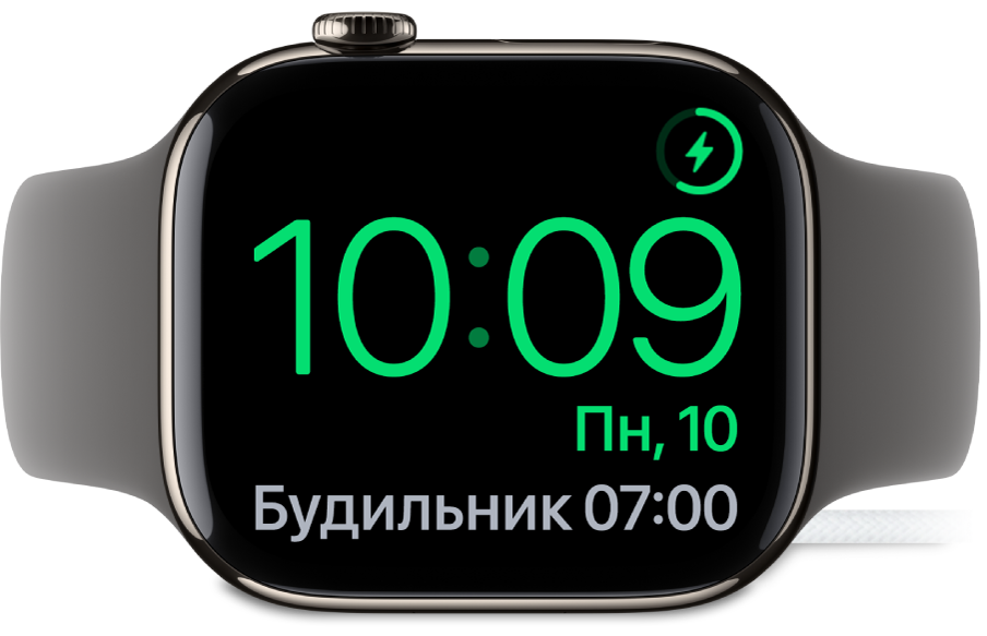 Apple Watch, поставленные набок и подсоединенные к зарядному устройству. На экране отображаются значок зарядки в правом верхнем углу, текущее время под ним и время следующего будильника.