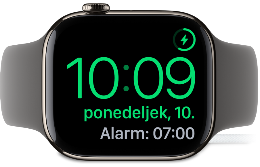 Ura Apple Watch na boku, priklopljena na polnilnik, na zaslonu pa je prikazan simbol za polnjenje v zgornjem desnem kotu, trenutni čas pod njim in čas naslednjega alarma.
