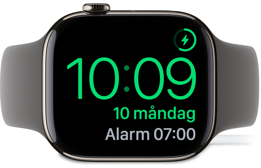 En Apple Watch som ligger på sidan och är ansluten till laddaren. Skärmen visar laddningssymbolen i det övre högra hörnet, under det tiden och sedan tiden för nästa alarm.
