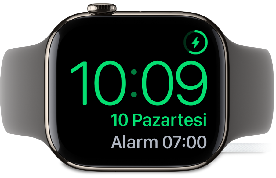 Sağ üst köşede şarj sembolünü, onun altında güncel saati ve bir sonraki alarmın saatini gösteren ekranıyla şarj aygıtına bağlı olarak yan yerleştirilmiş bir Apple Watch.