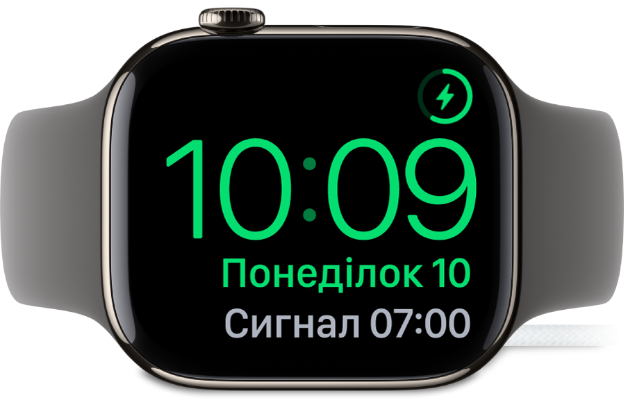 Apple Watch покладено на бік і підʼєднано до зарядного пристрою. На екрані відображається значок заряджання у верхньому правому куті, під ним — поточний час і час наступного будильника.