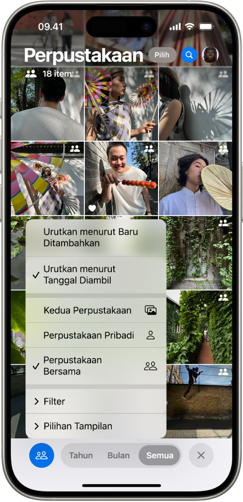 Perpustakaan Foto Bersama iCloud di app Foto. Tombol Perpustakaan Bersama di pojok kiri bawah dipilih, dan Perpustakaan Bersama dipilih di daftar opsi perpustakaan. Semua foto yang ditampilkan di perpustakaan memiliki tanda perpustakaan bersama di pojok kanan atas.