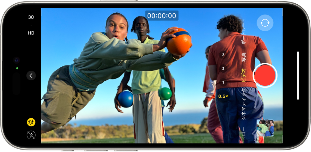 横向きのiPhone。「カメラ」が「ビデオ」モードで開いています。カメラの枠内には、ジョギング中の人が映っています。画面左下隅にはフラッシュボタンがあります。画面左側の中央にはカメラコントロールボタンがあります。左上隅にはビデオの解像度とフレームレートを切り替えるクイック切り替え機能があります。左上隅のアクションボタンがオンになっています。右側には、上から順に、背面カメラ選択ボタン、録画ボタン、写真およびビデオビューアボタンがあります。ビューファインダーに走っている人が映っています。