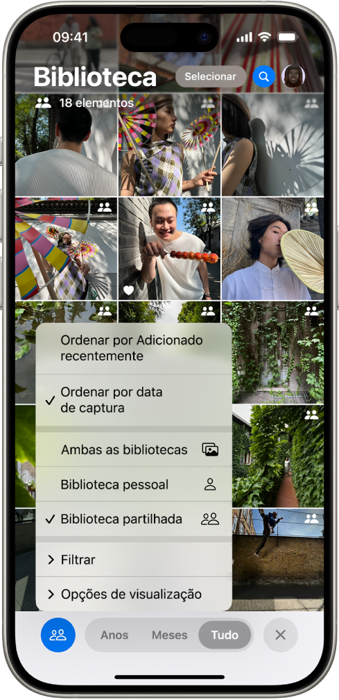 Uma fototeca partilhada em iCloud na aplicação Fotografias. O botão “Biblioteca partilhada” no canto inferior esquerdo está selecionado e “Biblioteca partilhada” está selecionada numa lista de opções da biblioteca. Todas as fotografias apresentadas na biblioteca têm um emblema de biblioteca partilhada no canto superior direito.