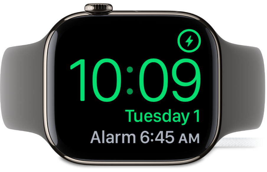 Apple Watch موضوعة على جانبها ومتصلة بالشاحن، وتعرض الشاشة رمز الشحن في الزاوية العلوية اليمنى، والوقت الحالي أسفل ذلك، ووقت المنبه التالي.