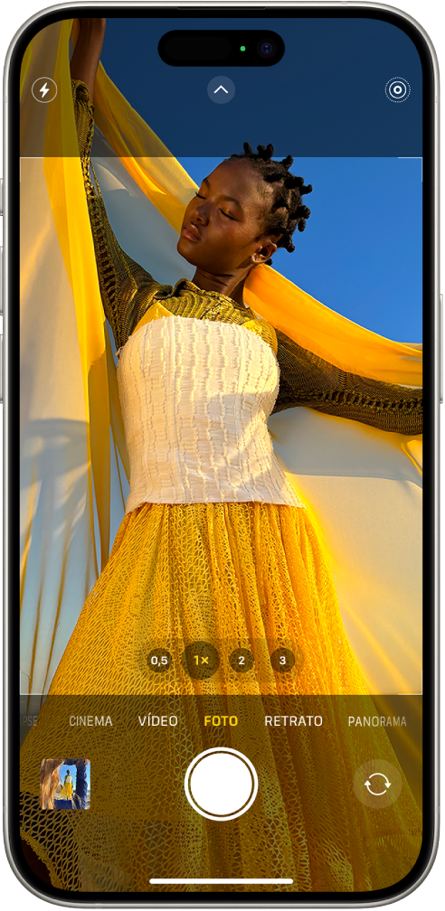 Câmera no modo Foto, com outros modos à direita e à esquerda, abaixo do visor. Os botões do Flash, Controles da Câmera e Live Photos aparecem na parte superior da tela. O botão Visualizador de Fotos e Vídeos está no canto inferior esquerdo. O botão Tirar Foto na parte inferior central e o botão Seletor de Câmera Traseira está no canto inferior direito.