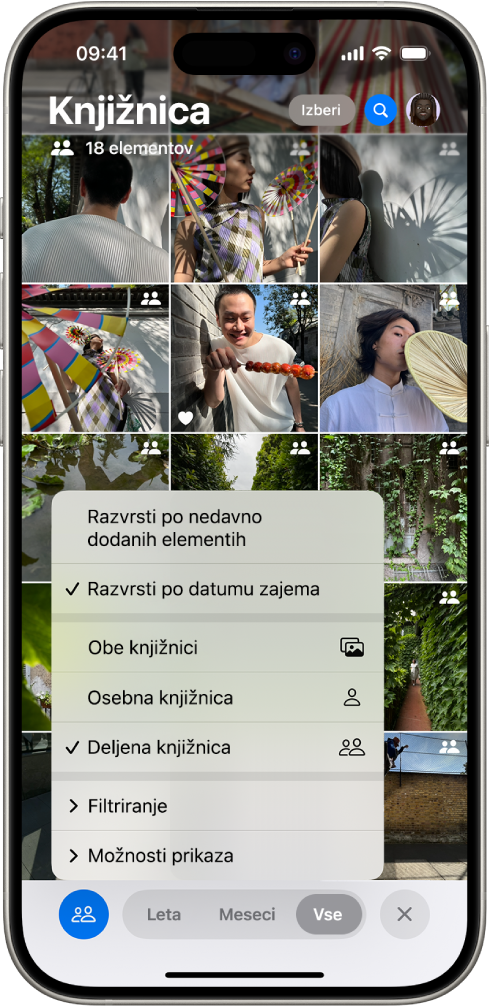 Deljena knjižnica fotografij v storitvi iCloud v aplikaciji Fotografije. Izbran je gumb Deljena knjižnica v spodnjem levem kotu in v seznamu možnosti knjižnice je izbrana Deljena knjižnica. Vse fotografije, prikazane v knjižnici, imajo v zgornjem desnem kotu oznako deljene knjižnice.
