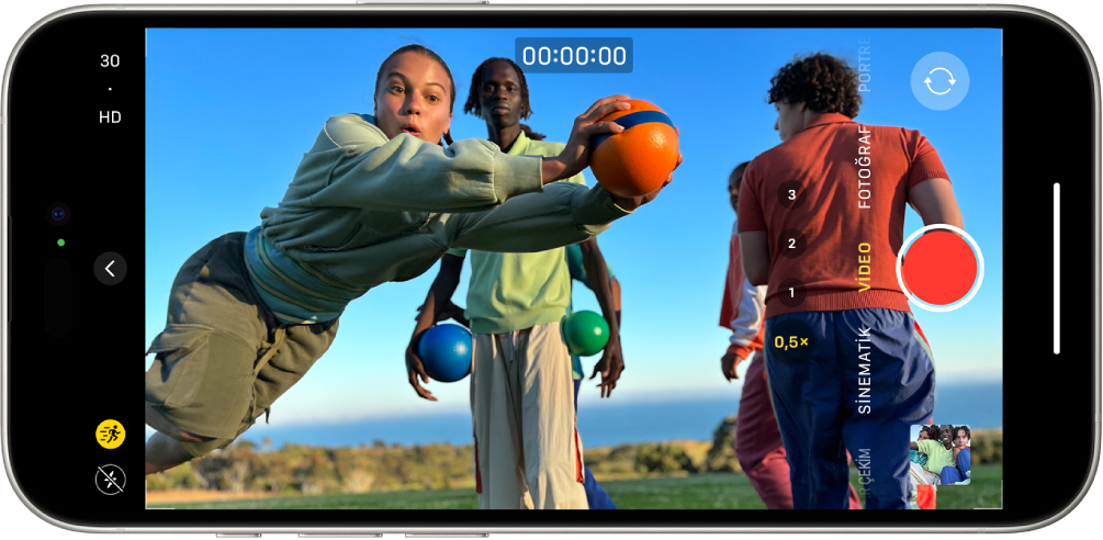 iPhone yatay olarak çevriliyor. Kamera açık ve Video modunda. Kamera çerçevesinde bir kişi koşuyor. El Feneri düğmesi ekranın sol alt köşesinde. Kamera Denetimleri düğmesi ekranın sol ortasında. Sol üst köşede, video çözünürlüğünü ve kare hızını değiştirmek için hızlı anahtarlar var. Sol üst köşedeki Hareket düğmesi açık. Sağ tarafta, en üstten en alta doğru Kamera Seçici Arka Yüz düğmesi, Kayıt düğmesi ve Fotoğraf ve Video Görüntüleyici düğmesi yer alıyor. Vizörün içinde bir kişi koşuyor.
