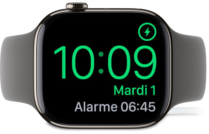 Apple Watch placée sur sa tranche et connectée au chargeur. L’écran montre le symbole de recharge dans le coin supérieur droit, l’heure actuelle en dessous et l’heure de la prochaine alarme.