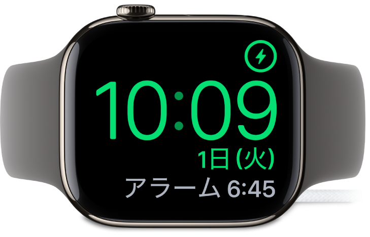 横向きに置かれ、充電器に接続されているApple Watch。画面には、右上隅に充電中のマーク、その下に現在時刻と、次のアラーム時刻が表示されています。