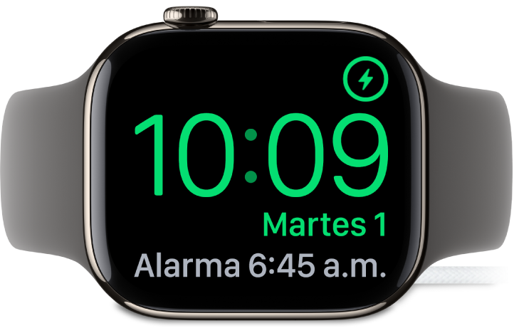 Un Apple Watch colocado de lado y conectado al cargador, con la pantalla mostrando el símbolo de carga en la esquina superior derecha, la hora actual debajo y la hora de la siguiente alarma.