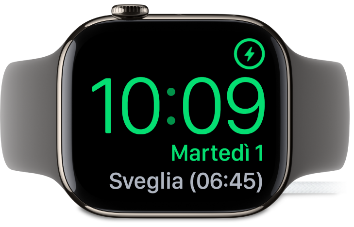 Apple Watch, appoggiato su un lato e collegato al caricabatterie, con lo schermo che mostra il simbolo di ricarica nell’angolo superiore destro, l’ora attuale sotto e l’orario della sveglia successiva.