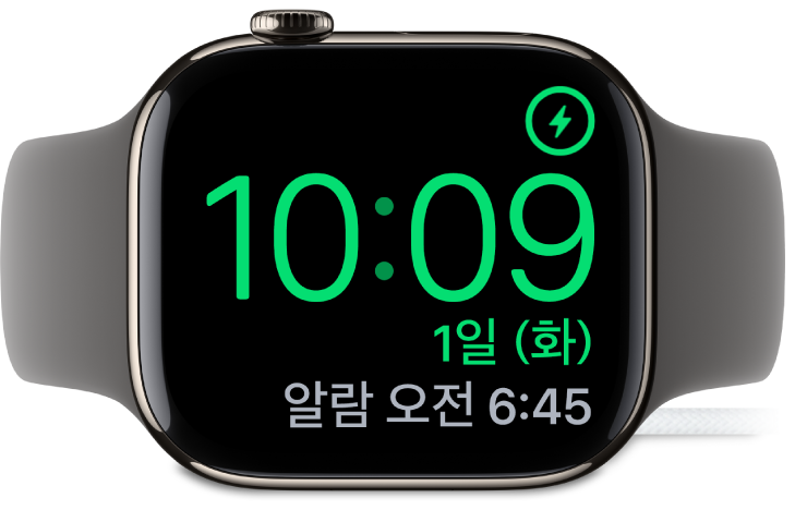 Apple Watch가 옆으로 누워 있고 충전기에 연결되어 있음. 화면의 오른쪽 상단 모서리에는 충전 기호가 있고 그 아래에는 현재 시간과 다음 알람 시간이 있음.