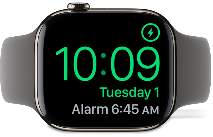 Apple Watch diletakkan di sisi dan bersambung ke pengecas, dengan skrin menunjukkan simbol mengecas di penjuru atas kanan, masa semasa di bawahnya dan masa penggera seterusnya.