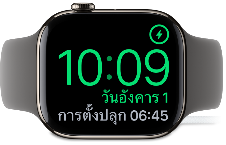 Apple Watch ที่วางตะแคง และเชื่อมต่ออยู่กับที่ชาร์จ โดยหน้าจอแสดงสัญลักษณ์การชาร์จตรงมุมขวาบนสุด เวลาปัจจุบันอยู่ด้านล่างสัญลักษณ์การชาร์จ และเวลาของการตั้งปลุกครั้งต่อไป