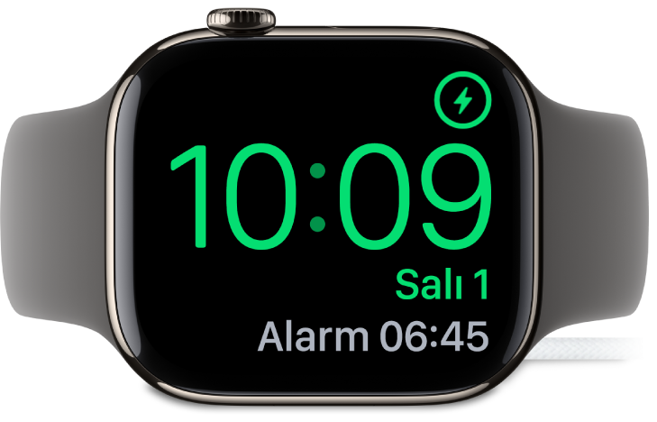 Sağ üst köşede şarj sembolünü, onun altında güncel saati ve bir sonraki alarmın saatini gösteren ekranıyla şarj aygıtına bağlı olarak yan yerleştirilmiş bir Apple Watch.