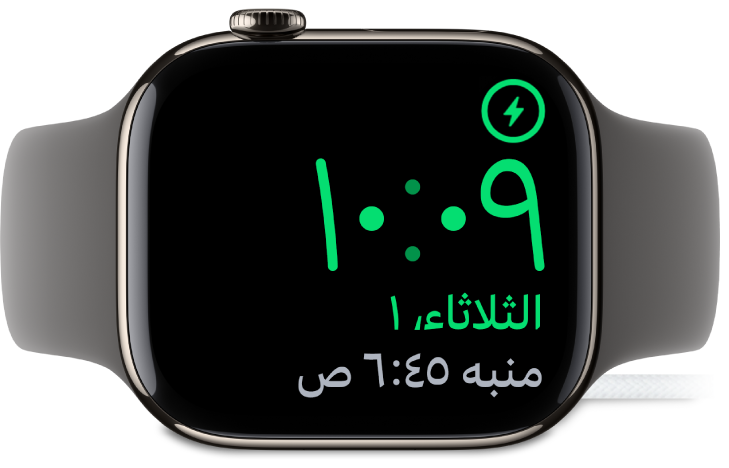 Apple Watch موضوعة على جانبها ومتصلة بالشاحن، وتعرض الشاشة رمز الشحن في الزاوية العلوية اليمنى، والوقت الحالي أسفل ذلك، ووقت المنبه التالي.