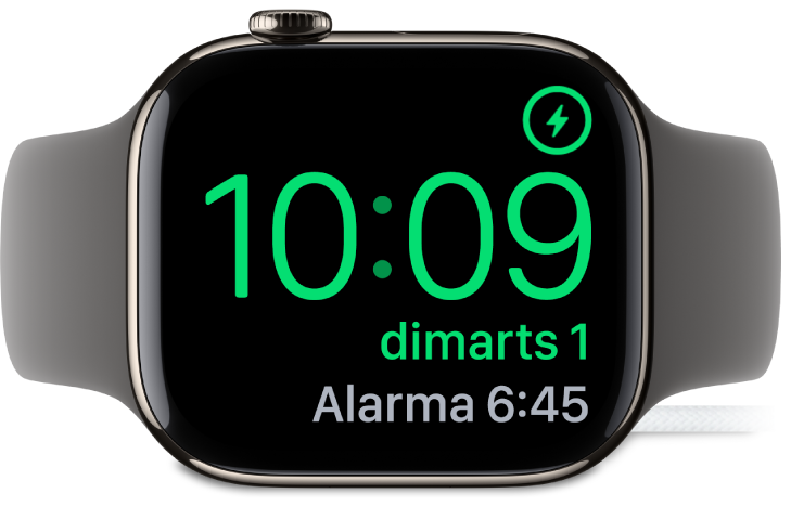 Un Apple Watch posat de costat i connectat al carregador amb la pantalla que mostra el símbol de càrrega a l’angle superior dret, l’hora actual a sota i l’hora de l’alarma següent.