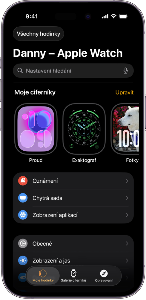Aplikace Apple Watch na iPhonu otevřená na obrazovce Moje hodinky. Nahoře se zobrazují ciferníky a pod nimi nastavení. Na dolním okraji obrazovky aplikace Apple Watch jsou tři panely: vlevo panel Moje hodinky, z kterého se Apple Watch nastavují; vedle něj panel Galerie ciferníků, na kterém si můžete prohlížet dostupné ciferníky a komplikace; a potom panel Objevování, kde se můžete o Apple Watch dozvědět víc