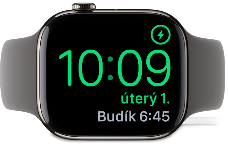 Hodinky Apple Watch položené na boku a připojené k nabíječce; na obrazovce se v pravém horním rohu ukazuje symbol nabíjení, pod ním aktuální čas a nejbližší nastavený čas buzení.