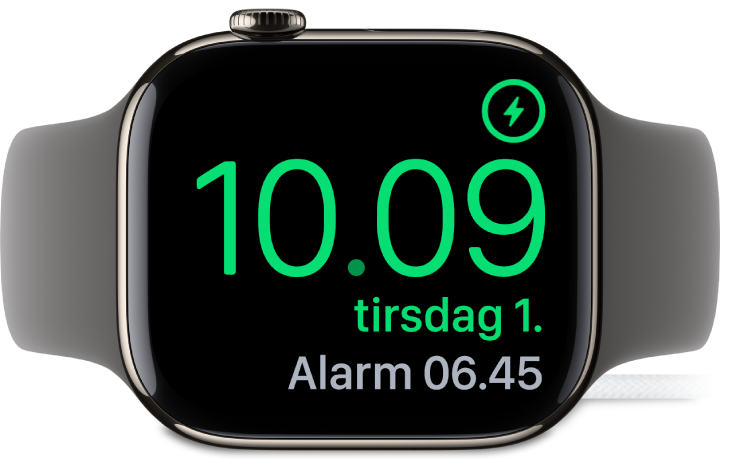 Et Apple Watch, der ligger på siden og er sluttet til opladeren, hvor skærmen viser opladersymbolet øverst til højre, det aktuelle klokkeslæt derunder og klokkeslættet for den næste alarm.