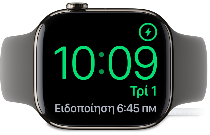 Ένα Apple Watch είναι σε πλάγια θέση και συνδεδεμένο στον φορτιστή του. Στην οθόνη εμφανίζονται το σύμβολο φόρτισης στην πάνω δεξιά γωνία, η τρέχουσα ώρα από κάτω, και η ώρα της επόμενης ειδοποίησης.