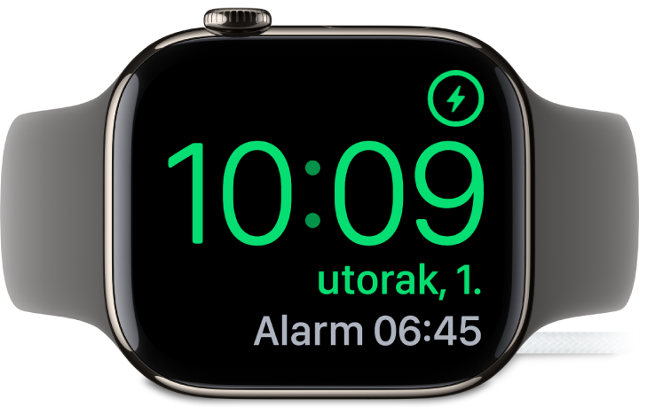 Apple Watch postavljen na bok i spojen na punjač, na čijem zaslonu je u gornjem desnom kutu prikazan simbol napajanja, trenutačno vrijeme dolje ispod i vrijeme sljedećeg alarma.