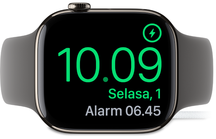 Apple Watch diletakkan di sampingnya dan tersambung ke pengisi daya, dengan layar menampilkan simbol pengisian daya di pojok kanan atas, waktu saat ini di bawahnya, dan waktu alarm berikutnya.