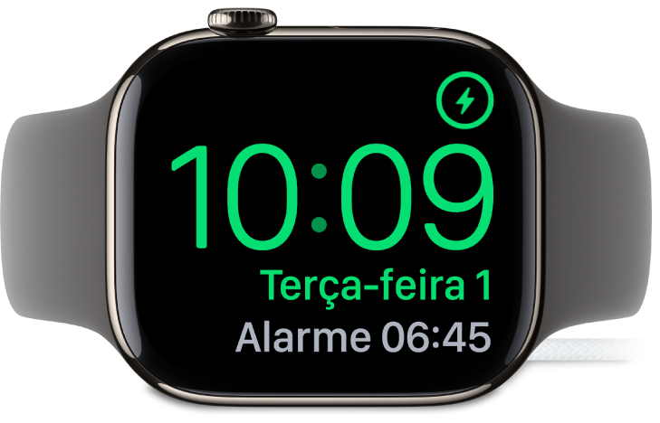 Apple Watch posicionado de lado e conectado ao carregador, com a tela mostrando o símbolo de carregamento no canto superior direito, a hora atual abaixo e o horário do próximo alarme.