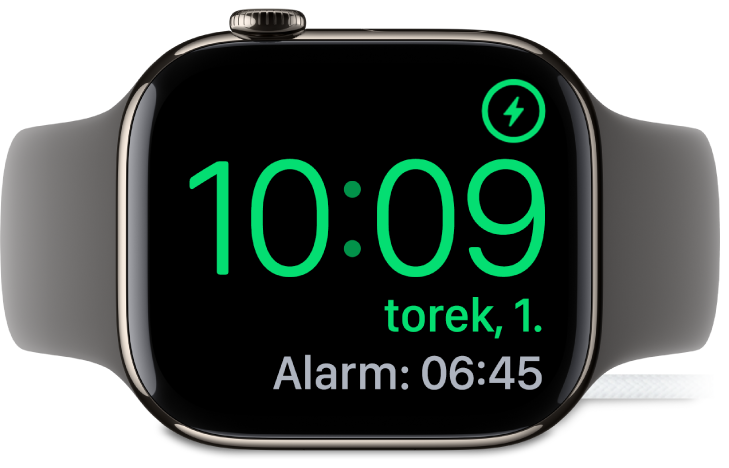 Ura Apple Watch na boku, priklopljena na polnilnik, na zaslonu pa je prikazan simbol za polnjenje v zgornjem desnem kotu, trenutni čas pod njim in čas naslednjega alarma.