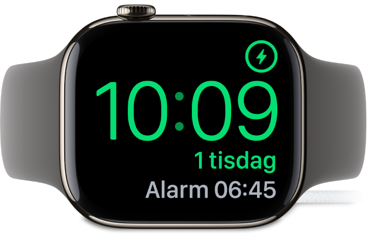 En Apple Watch som ligger på sidan och är ansluten till laddaren. Skärmen visar laddningssymbolen i det övre högra hörnet, under det tiden och sedan tiden för nästa alarm.