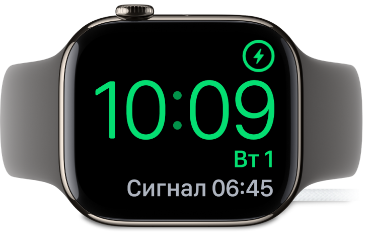 Apple Watch покладено на бік і підʼєднано до зарядного пристрою. На екрані відображається значок заряджання у верхньому правому куті, під ним — поточний час і час наступного будильника.
