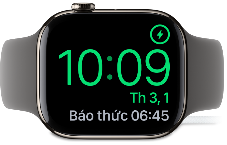 Một Apple Watch được đặt nghiêng và được kết nối với bộ sạc, với màn hình đang hiển thị biểu tượng đang sạc ở góc trên cùng bên phải, thời gian hiện tại ở bên dưới và thời gian của báo thức tiếp theo.