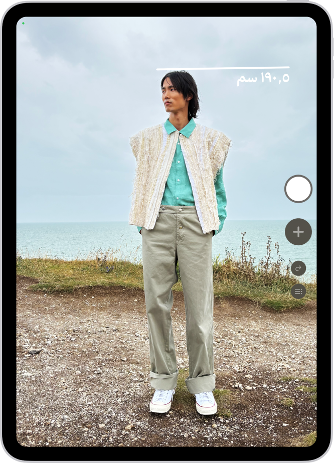 شخص يتم قياس طوله في تطبيق القياس، ويظهر قياس الطول أعلى رأس الشخص. زر التقاط صورة بالقرب من الزاوية السفلية اليسرى لالتقاط صورة للقياس. ويظهر مؤشر الكاميرا المستخدمة الأخضر في الأعلى.