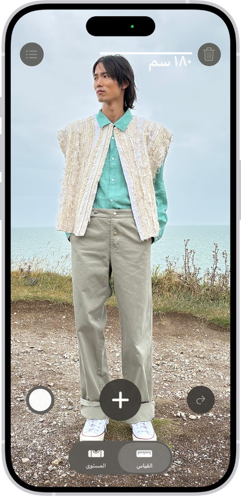 شخص يتم قياس طوله في تطبيق القياس، ويظهر قياس الطول أعلى رأس الشخص. زر التقاط صورة بالقرب من الزاوية السفلية اليسرى لالتقاط صورة للقياس. ويظهر مؤشر الكاميرا المستخدمة الأخضر في الأعلى.